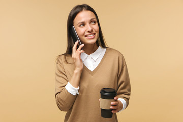 Wall Mural - Studio portrait of businesswoman isolated on brown background, holding takeaway coffee cup while talking on phone, smiling positively and looking sideways as if waiting