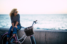 Happy Beautiful Curly Healthy Caucasian Adult Middle Age Woman Sit Down On A Vintage Coloured Bike And Enjoy The Ocean In Outdoor Leisure Activity - Trendy People With Hapy Life