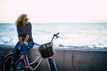 Happy beautiful curly healthy caucasian adult middle age woman sit down on a vintage coloured bike and enjoy the ocean in outdoor leisure activity - trendy people with hapy life