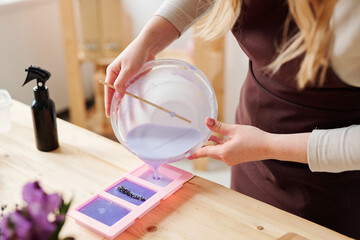 Hands of craftswoman in apron pouring lilac liquid soap mass into silicone molds