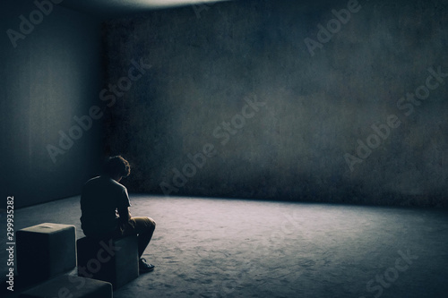 Fotografía Solitary depressed lonely sad man sitting on his own alone in large empty dark room with plain blank wall copy space seated with head bowed down behind spotlight