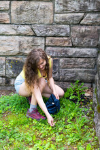 Young Woman Happy Girl Foraging Picking Wild Green Dandelion Leaves For Health In Park By Wooden Wall Corner Bending Over Crouching Down