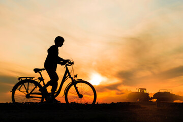 Boy , kid 10 years old riding bike in countryside, tractor working in background,  silhouette of riding person and machine at sunset in nature