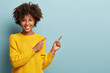 canvas print picture - Cheerful Afro woman points away on copy space, discusses amazing promo, gives way or direction, wears yellow warm sweater, has pleasant smile, feels optimistic, isolated over blue background