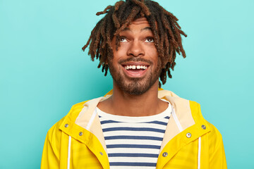 Wall Mural - Headshot of happy man with dreads, has positive expression, focused above, wears sailor jumper and yellow raincoat, isolated over blue background, enjoys rainy autumn weather. Cheerful teenager