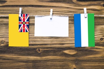 Hanging flags of Niue and Sierra Leone attached to rope with clothes pins with copy space on white note paper on wooden background.Diplomatic relations between countries.