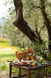 © ArtSys - Romantic picnic under olive tree. Delicious italian meal served on a wooden table. Baskets with food, branches in glass jar. Sunny autumn day. Italy, Tuscany