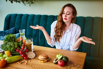 Wall Mural - Beautiful young woman decides eating hamburger or vegetables  in kitchen. Cheap junk food vs healthy diet