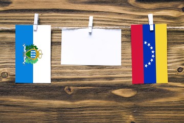 Hanging flags of San Marino and Venezuela attached to rope with clothes pins with copy space on white note paper on wooden background.Diplomatic relations between countries.