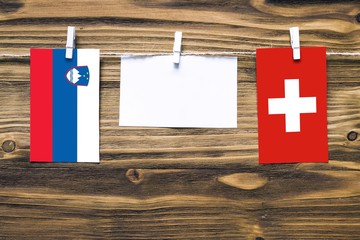 Hanging flags of Slovenia and Switzerland attached to rope with clothes pins with copy space on white note paper on wooden background.Diplomatic relations between countries.