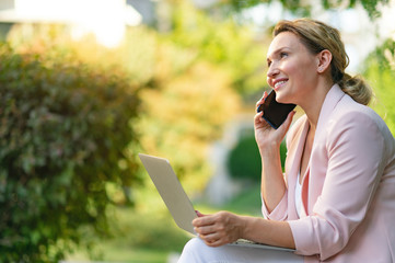 Close-up portrait of a smiling woman calling by phone on the street.  Happy businesswoman is using smart phone, outdoors. Cheerful businesswoman in a jacket with cell phone in park.