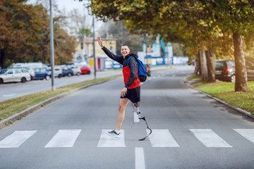 Wall Mural - Full length of handsome caucasian handicapped sportsman in sportswear, with artificial leg and backpack crossing street and waving to a friend.