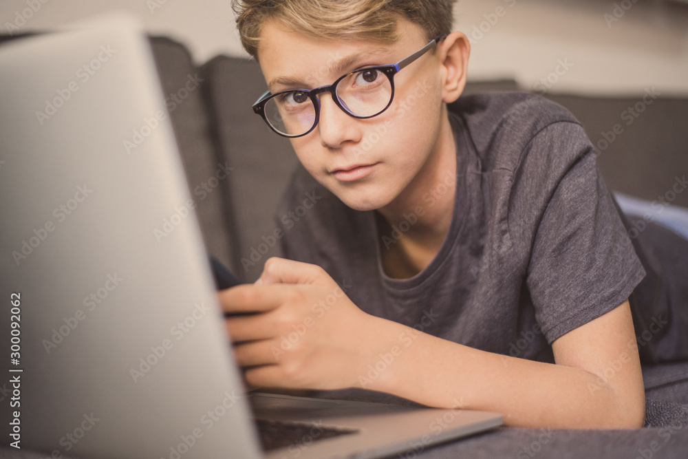 Young student doing homework lying on a sofa using laptop and smartphone. Caucasian boy communicate with friends. Teen enjoying  new electronic devices. Technology education concept Soft focus on eyes - obrazy, fototapety, plakaty 