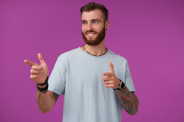 Positive pretty tattooed brunette guy with trendy haircut wearing blue t-shirt, pointing aside with forefingers and smiling widely, posing over purple background