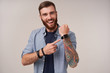 Joyful young pretty bearded brunette man with tattooed hands smiling widely to camera and pointing to his watch, being in high spirit while posing over white background