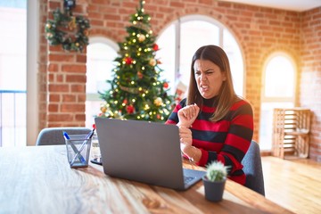 Poster - Beautiful woman sitting at the table working with laptop at home around christmas tree disgusted expression, displeased and fearful doing disgust face because aversion reaction. With hands raised