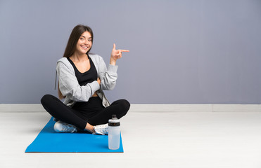 Wall Mural - Young sport woman sitting on the floor with mat pointing finger to the side