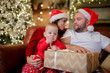 © Sergey Mironov - Image of young parents in Santa's caps with son sitting in chair in room .