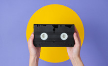 Female Hand Holding Video Cassette On Purple Background With Yellow Circle