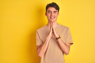 Wall Mural - Teenager boy wearing yellow t-shirt over isolated background praying with hands together asking for forgiveness smiling confident.