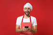 Excited young bearded male chef cook or baker man in striped apron toque chefs hat isolated on bright red background. Cooking food concept. Mock up copy space. Using mobile phone, typing sms message.