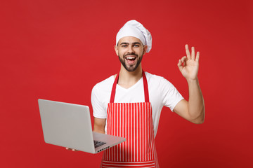Funny young bearded chef cook or baker man in striped apron t-shirt toque chefs hat isolated on red background. Cooking food concept. Mock up copy space. Hold laptop pc computer, showing OK gesture.