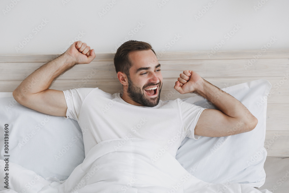 Calm young handsome bearded man lying in bed with white sheet pillow blanket in bedroom at home. Smiling beauty male spending time in room. Rest relax good mood lifestyle concept. Mock up copy space. - obrazy, fototapety, plakaty 