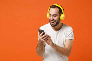 Canvas Print - Funny young man in casual white t-shirt posing isolated on orange wall background studio portrait. People lifestyle concept. Mock up copy space. Listening music with headphones, hold mobile phone.