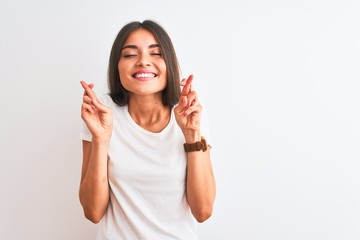 Wall Mural - Young beautiful woman wearing casual t-shirt standing over isolated white background gesturing finger crossed smiling with hope and eyes closed. Luck and superstitious concept.