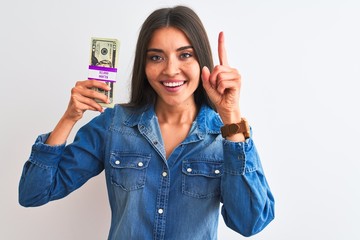 Wall Mural - Young beautiful woman holding dollars standing over isolated white background surprised with an idea or question pointing finger with happy face, number one