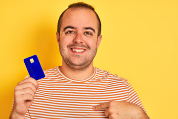 Sticker - Young man holding blue credit card standing over isolated yellow background with surprise face pointing finger to himself