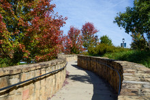 Looking Up A Winding Walkway. Framed By A Rock Wall, The Sidewalk Is Surround By Fall Colored Trees.