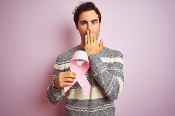 Poster - Young handsome man holding cancer ribbon standing over isolated pink background cover mouth with hand shocked with shame for mistake, expression of fear, scared in silence, secret concept