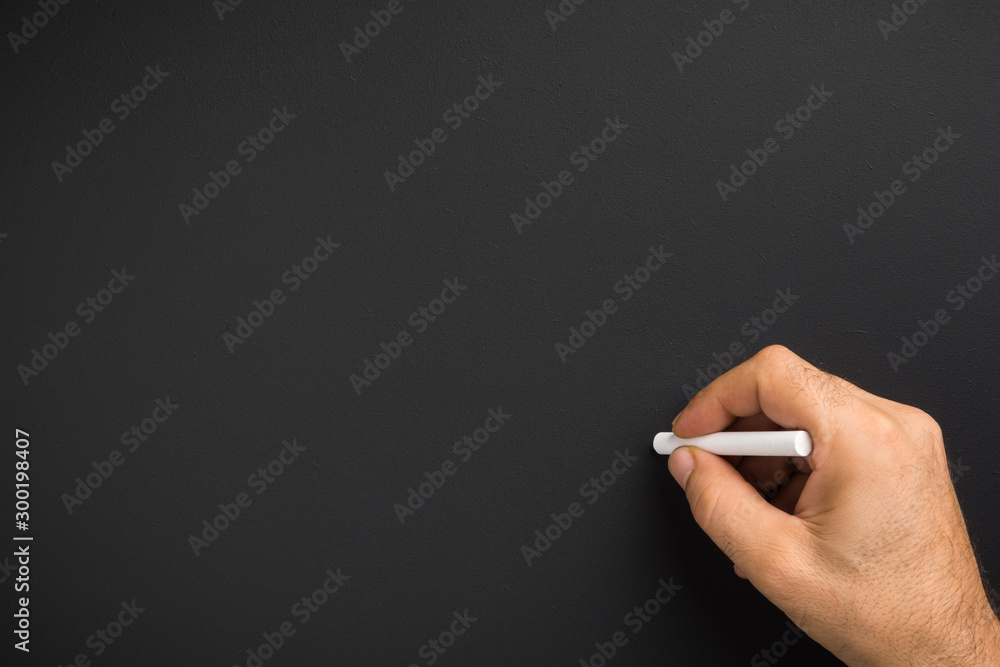 Close up of man hand holding chalk against blank black chalkboard - obrazy, fototapety, plakaty 