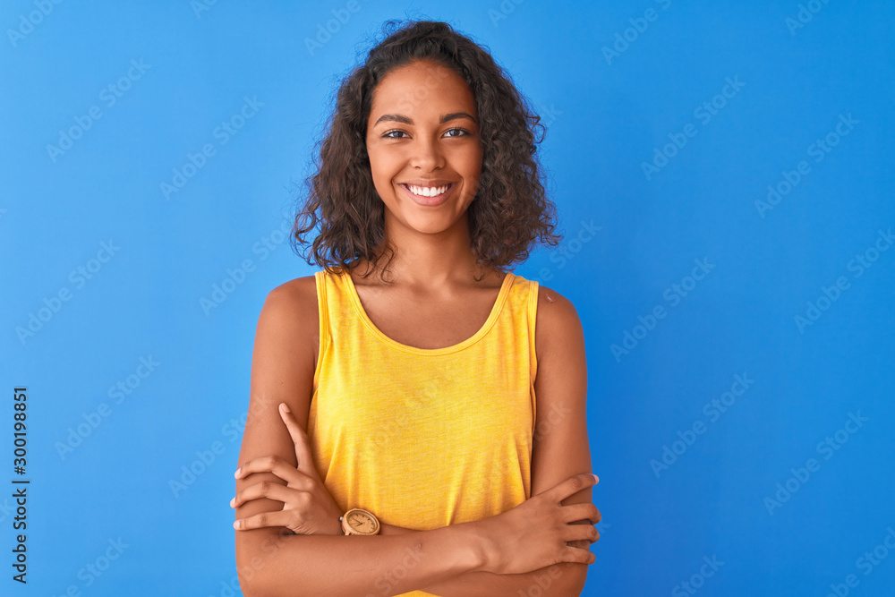 Young brazilian woman wearing yellow t-shirt standing over isolated blue background happy face smiling with crossed arms looking at the camera. Positive person.