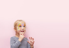 Pretty Exciting Smiling Blond Little Child Girl Of 3 Years With A Bright Face Painting On Pink Background. Fun Emotions, Happy Childhood. Holds A Finger Near His Mouth, Thinks And Looks Up