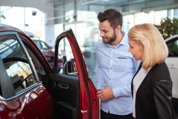 Wall Mural - Handsome man and his wife looking inside a new car for sale at the dealership, copy space. Couple buying new car together