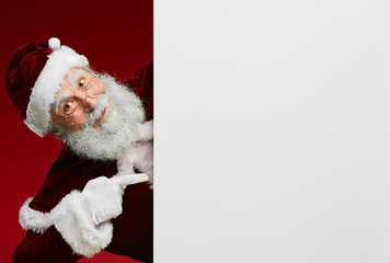 Wall Mural - Portrait of classic Santa Claus smiling gleefully at camera while pointing at blank white sign, copy space