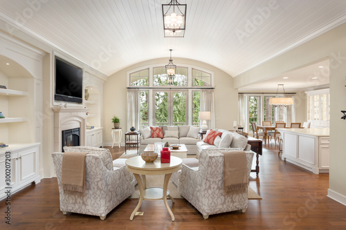 Living room in traditional style luxury home, with view of kitchen and dining area. Features tall ceilings, fireplace with tv, and French doors leading outside.