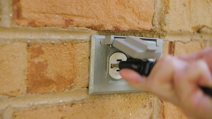 Wall Mural - Man plugging in a power cord and then unplugging it from an outdoor electrical outlet. Dual covered outdoor power sockets on brick wall. Black extension cord plug inserted into a wall socket.