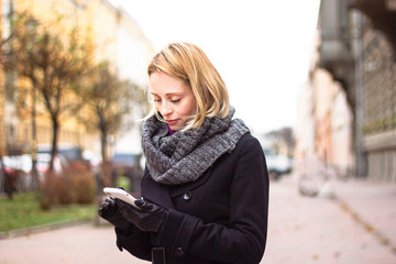 Girl with a phone in the autumn park.  Woman on a cloudy day dressed in seasonal weather, looking at her cell phone. Young blonde lady is typing a text message on smartphone