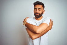 Young Indian Man Wearing T-shirt Standing Over Isolated White Background Hugging Oneself Happy And Positive, Smiling Confident. Self Love And Self Care