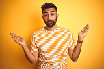 Wall Mural - Young indian man wearing t-shirt standing over isolated yellow background clueless and confused expression with arms and hands raised. Doubt concept.