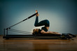 © Georgii - Young girl doing pilates exercises with a reformer bed. Beautiful slim fitness trainer on reformer gray background, low key, art light. Fitness concept