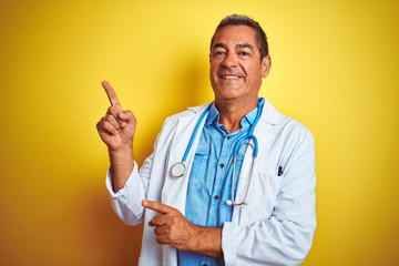 Wall Mural - Handsome middle age doctor man wearing stethoscope over isolated yellow background smiling and looking at the camera pointing with two hands and fingers to the side.