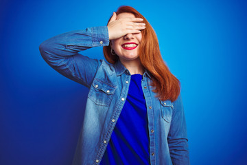 Canvas Print - Young beautiful redhead woman wearing denim shirt standing over blue isolated background smiling and laughing with hand on face covering eyes for surprise. Blind concept.