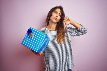 Wall Mural - Young beautiful woman holding birthday gift over pink isolated background with angry face, negative sign showing dislike with thumbs down, rejection concept