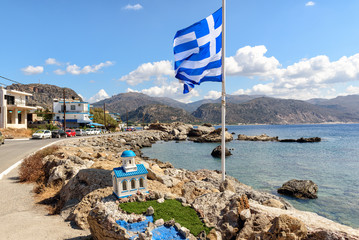 Big blue Greek flag and small church at coast of Palaiochora town on Crete island, Greece