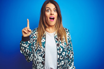 Canvas Print - Young beautiful woman wearing a jacket standing over blue isolated background pointing finger up with successful idea. Exited and happy. Number one.