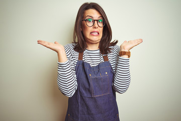 Poster - Young beautiful business woman wearing store uniform apron over isolated background clueless and confused expression with arms and hands raised. Doubt concept.